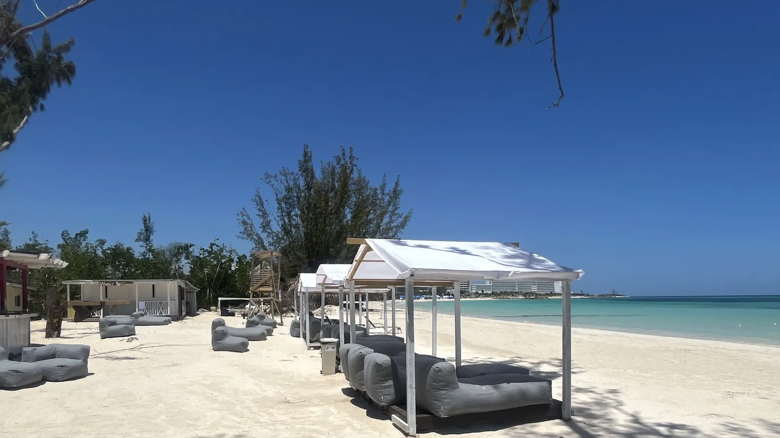 A white-canopied cabana with grey daybeds on the raked white sand at 876 Beach Club, turquoise shallows to the right, a lifeguard tower and water sports hut behind, and a large resort on the far point