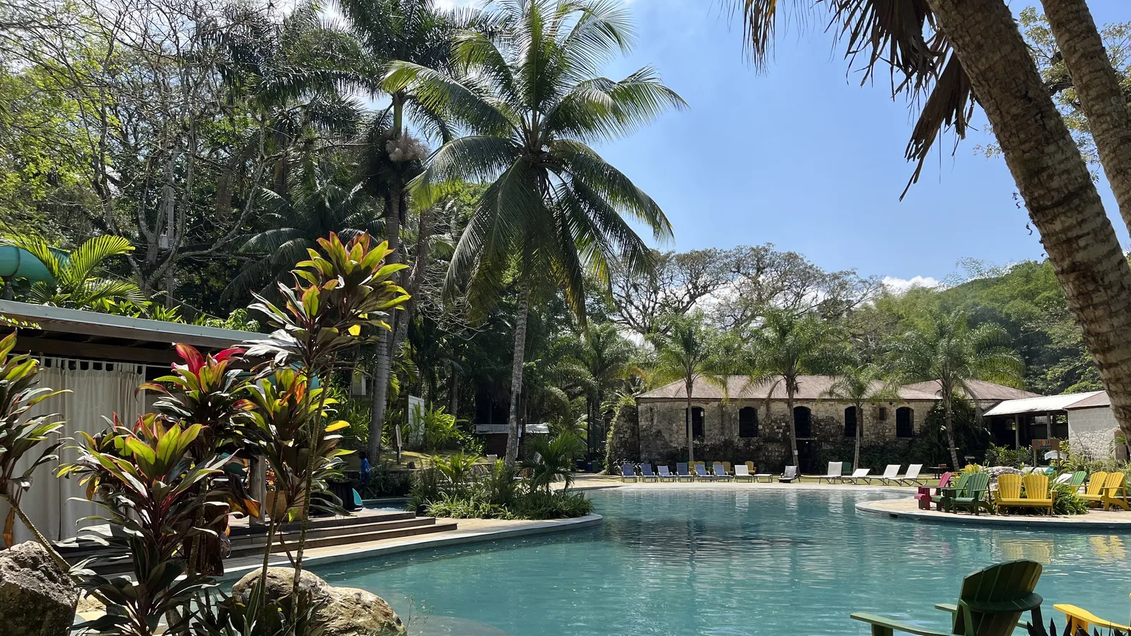 The freeform pool at Chukka's Good Hope outpost, ringed by palms and tropical planting, with bright Adirondack chairs on the far deck and the estate's old cut-stone works building behind