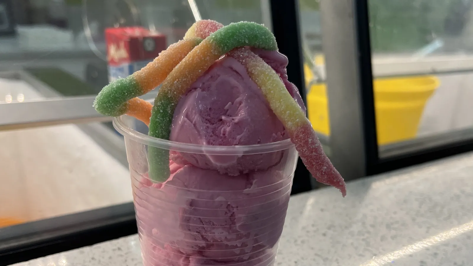A clear cup of bright pink ice cream topped with sugared sour gummy worms, on the counter at Devon House I Scream