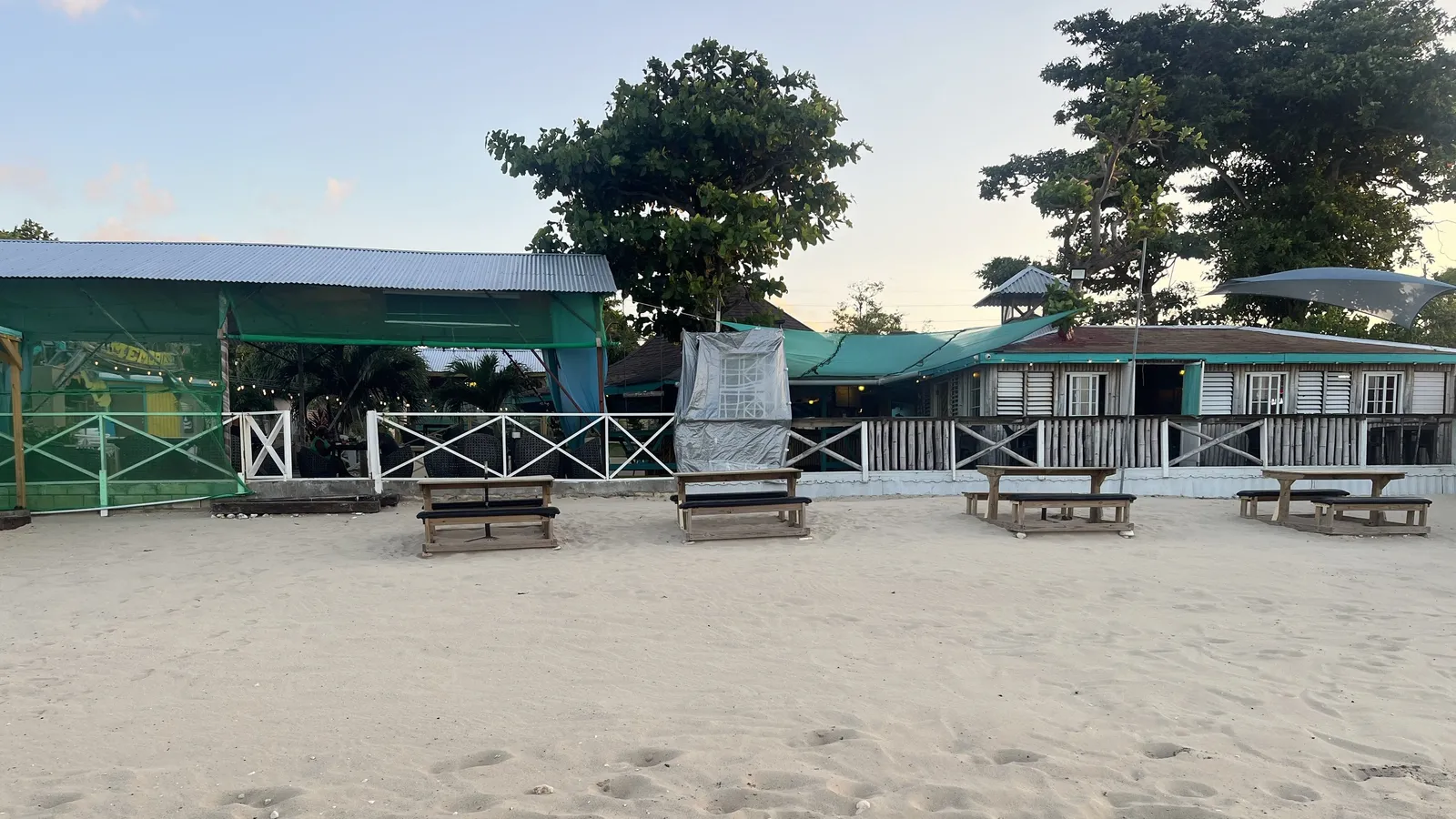 TikiTail's beachfront bar at dusk — a wooden building with turquoise shutters and string lights behind a low white fence, with picnic benches set out on the sand